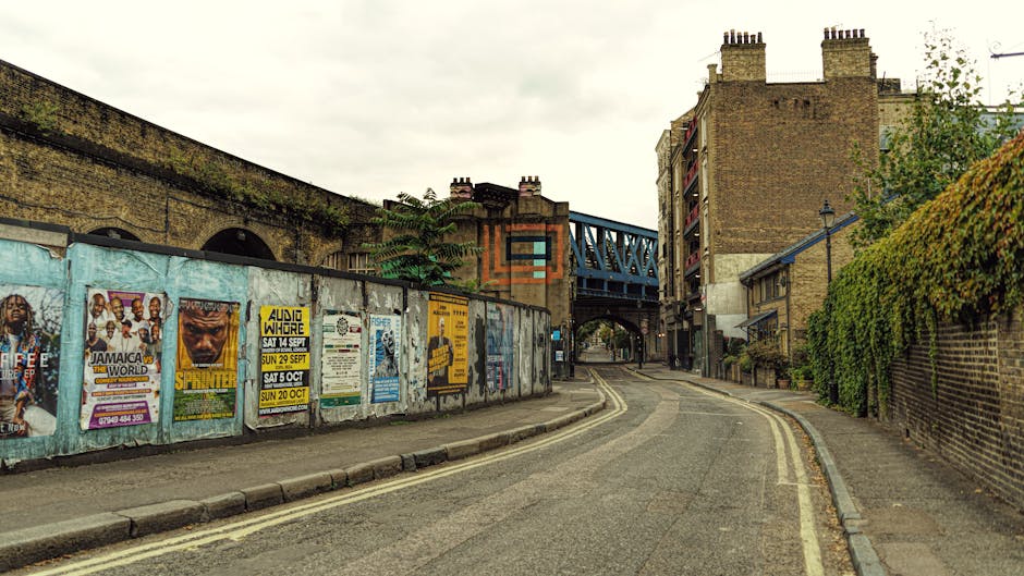 A busy street scene in Kentish Town showing a line of parked cars and delivery vans close to the pavement, with pedestrians walking past. Several brick terraced buildings with large windows line the s