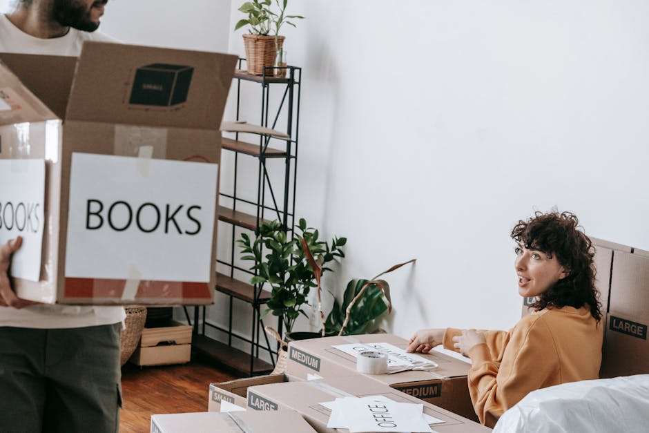 A man and a woman in a room with natural light from a window, surrounded by numerous packed cardboard boxes of various sizes, some labeled with handwritten descriptions such as 'books'. The woman is s