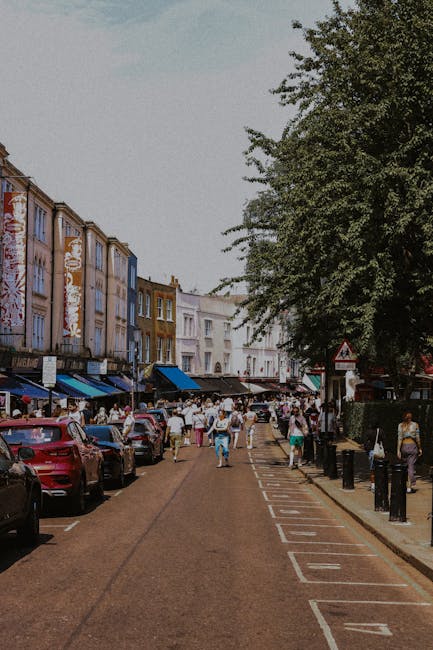 A busy street scene in Kentish Town with a row of multicoloured terraced houses on the left, some of which feature commercial signage and storefronts with awnings. The street is lined with parked cars