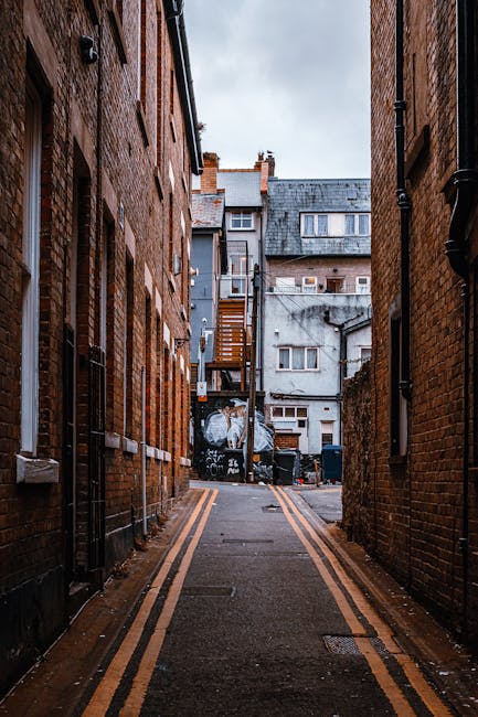A narrow alleyway between two tall brick buildings in an urban residential area, with a paved surface marked by double yellow lines along each side. At the end of the alley, there are several residential structures, including a grey building with multiple small windows and a wooden staircase attached to one of them, leading to an upper floor. The alley contains various items associated with a home relocation process, such as large black trash bins, plastic-wrapped furniture, and cardboard boxes stacked near the buildings, suggesting packing and moving activities. The scene is lit by natural daylight, with an overcast sky overhead. The image, provided by Removal Company Kentish Town, captures the environment where furniture transport and loading may occur during house removals or flat moves, reflecting the logistical aspects of a professional moving service on Fortess Road.