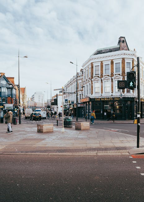 A busy street scene in Kentish Town showing a row of commercial buildings, including a pharmacy and beauty shop, with large retail windows and signs. Several pedestrians are walking along the pavement